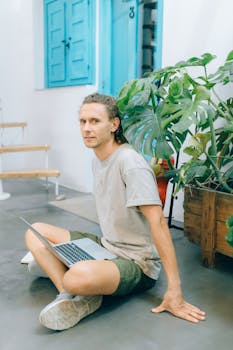 Man sitting on a floor with a laptop amidst indoor plants, showcasing a relaxed workspace.