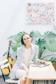 Woman in a blazer with eyeglasses smiling indoors at a desk. Modern and vibrant setting.