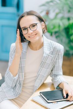 办公室里，一位穿着西装外套、戴眼镜的女人拿着笔记本，在办公桌前微笑