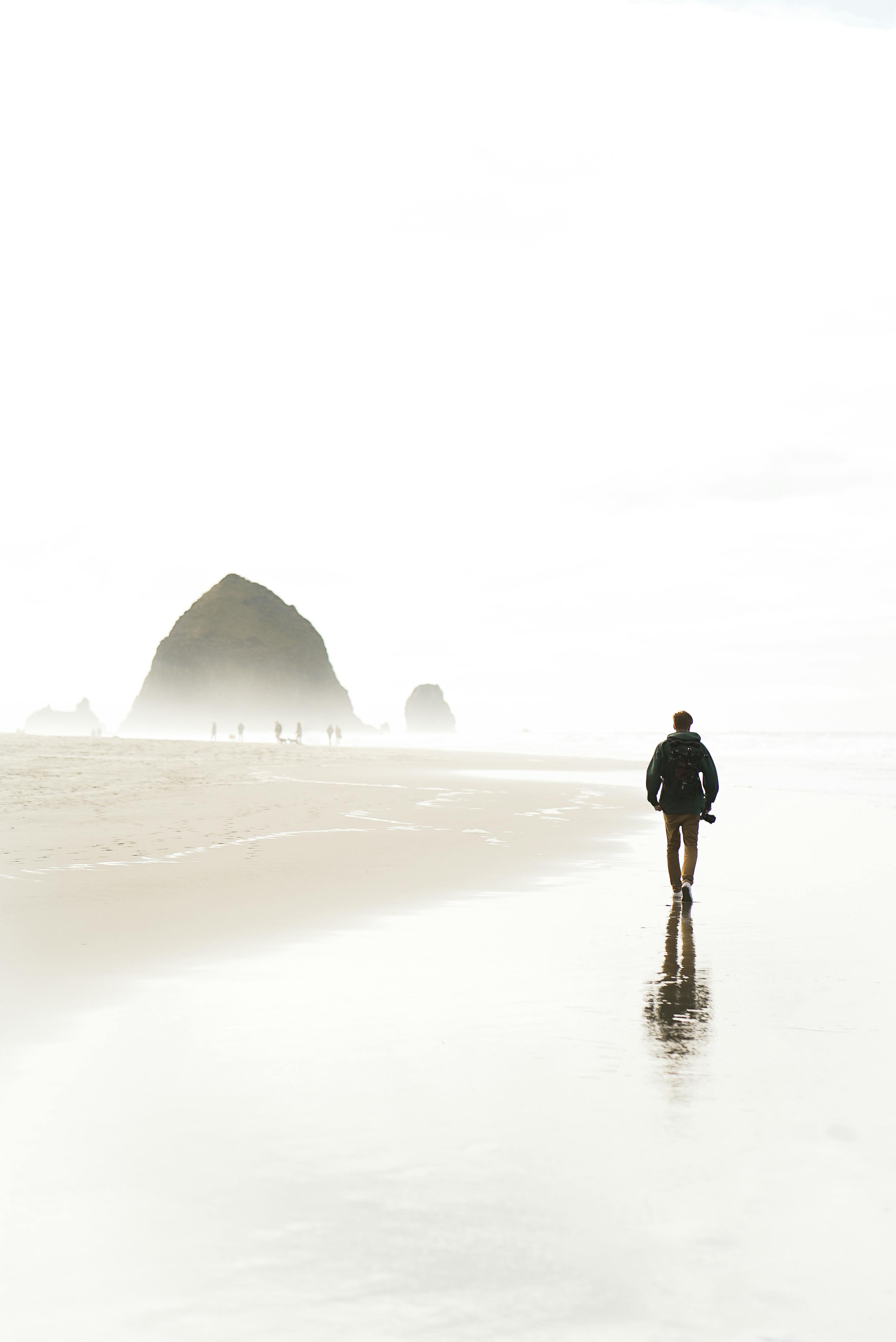 Back view of anonymous male tourist in casual outfit walking on wet sandy beach of ocean near rocky formations