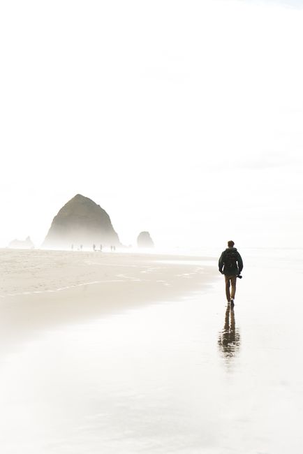 Back view of anonymous male tourist in casual outfit walking on wet sandy beach of ocean near rocky formations