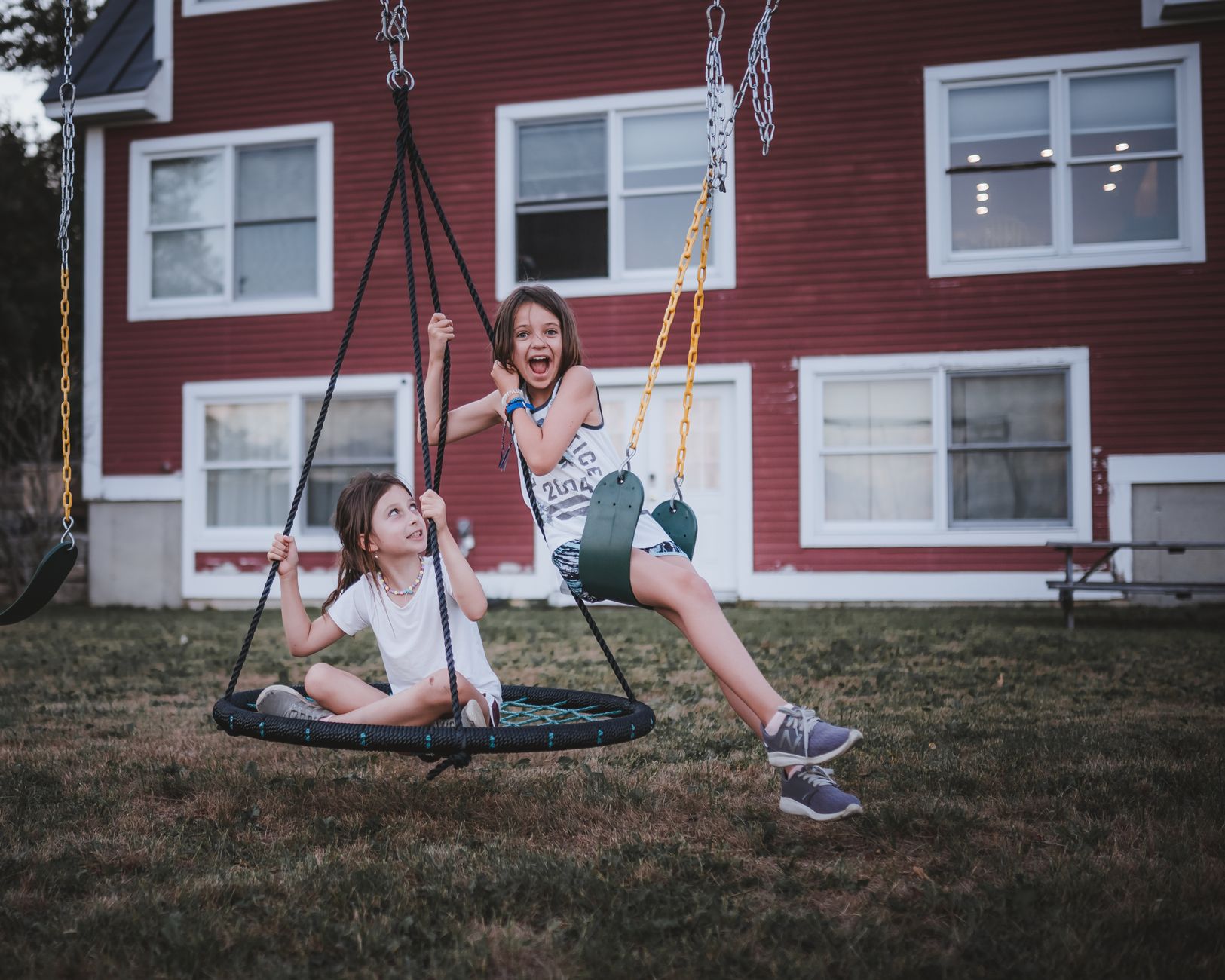Full length of positive children in casual clothes sitting on swing on playground on grassy lawn near red building in summer evening