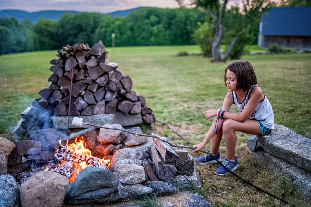 Full body of little girl in summer outfit sitting on stone bench and roasting marshmallow on twig on fire near boulders and logs on grassy field near trees in summer evening