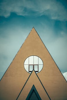 A modern triangular building facade with circular window against a cloudy sky, showcasing minimalist design.