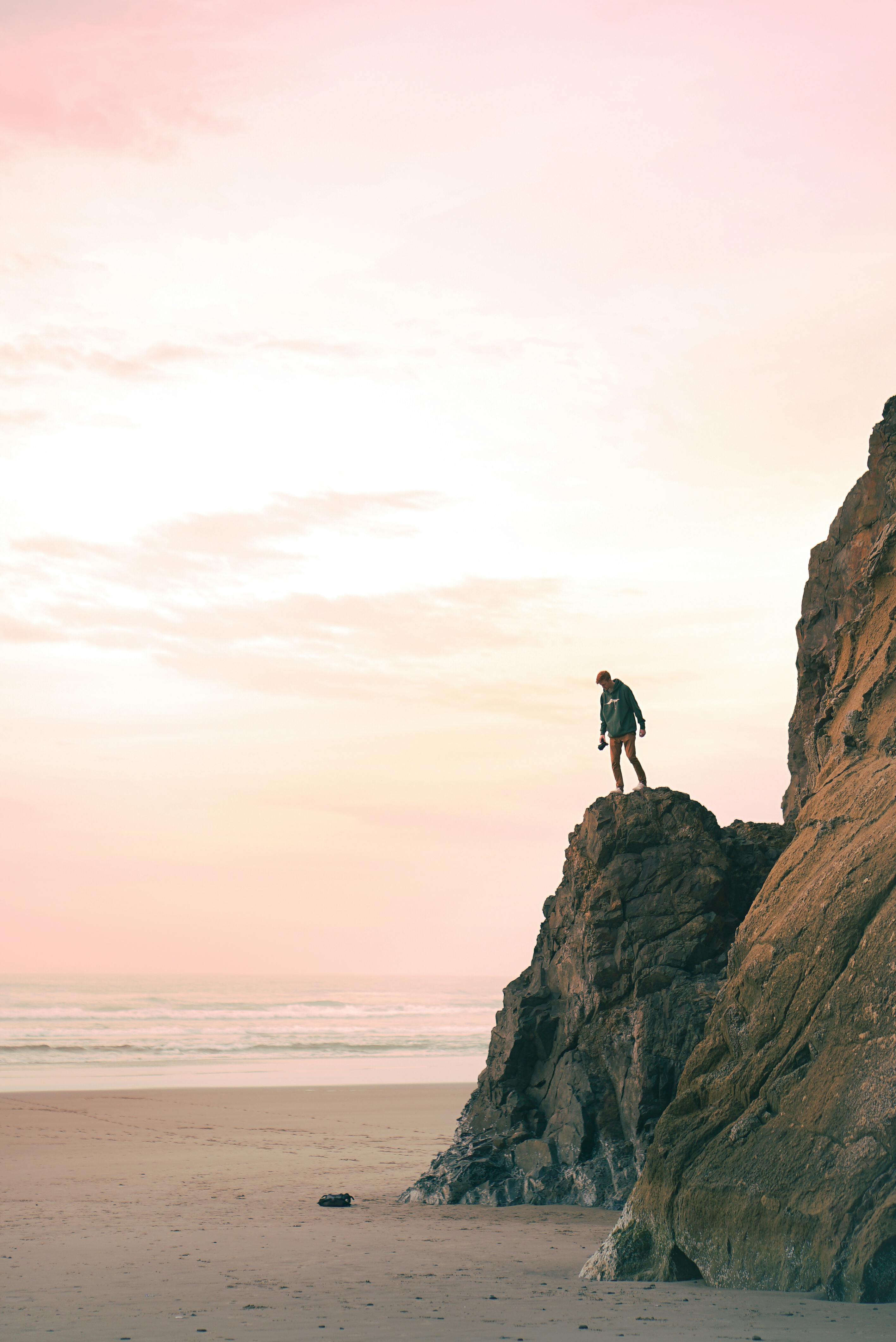 Distant unrecognizable tourist standing on rough rocky cliff located on sandy beach and admiring spectacular view of sunset over ocean