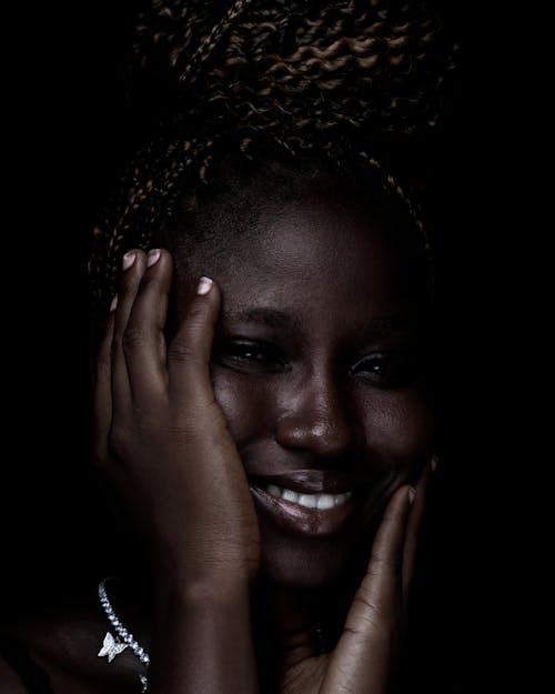 Young African female touching face with hands while smiling brightly at camera at black background