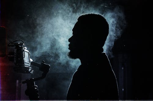 Side view silhouette of serious bearded male videographer recording video on professional video camera while standing in dark smoky studio