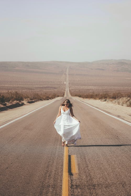 Full body of anonymous young female in maxi white dress walking on empty road among dry fields in semi desert countryside
