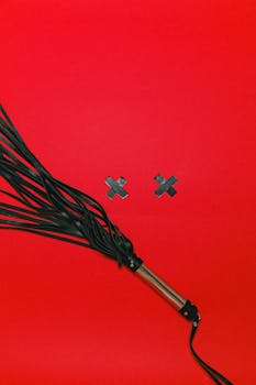 Black leather whip with tape crosses on a vibrant red background, conveying sensuality.
