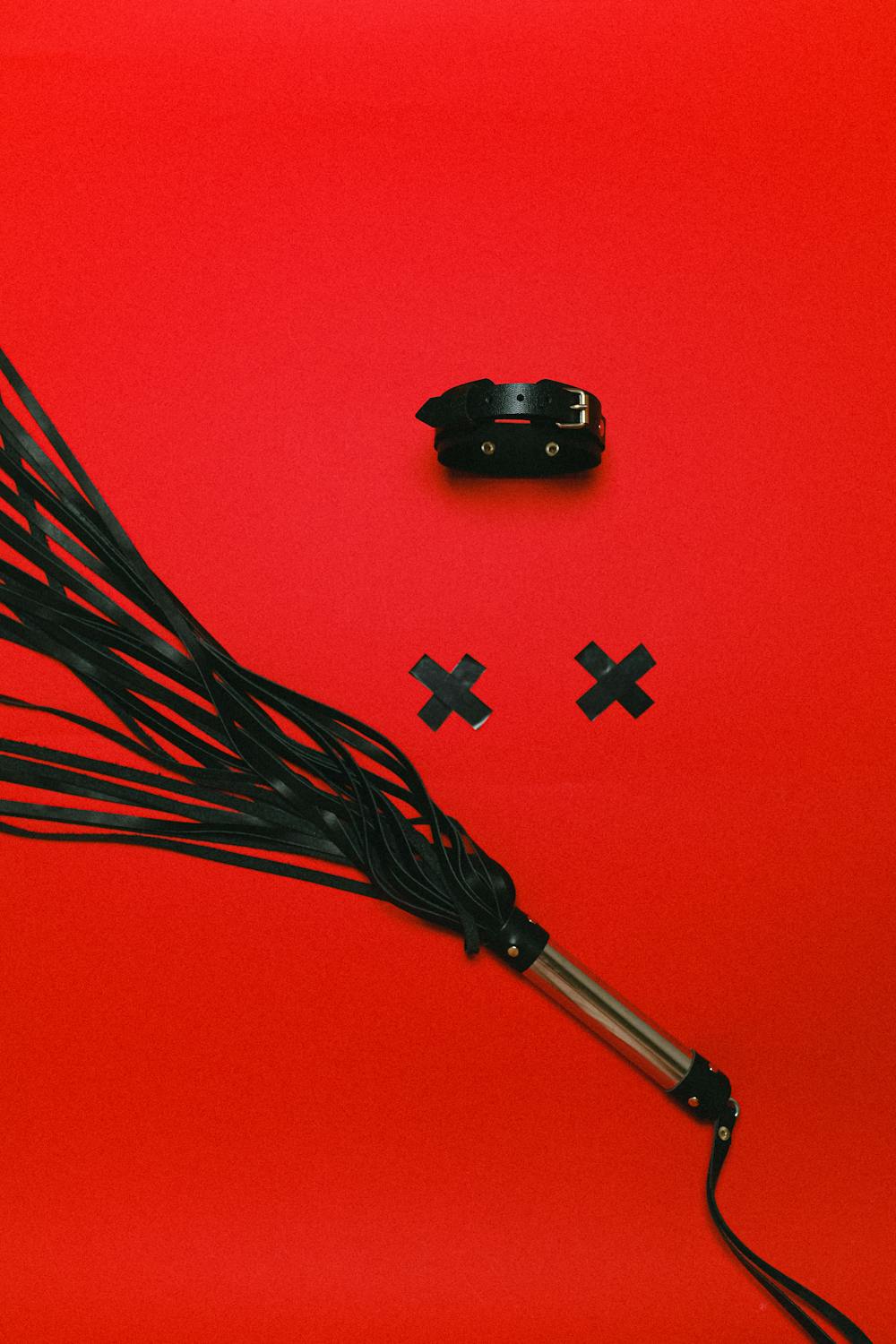 A picture of a black leather collar, two Xs made of electrical tape, and a black leather flogger on a bright red background.