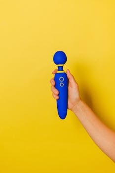 A blue vibrator held by a hand on a bright yellow backdrop.