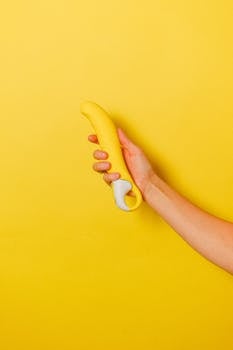 A hand holding a yellow silicone vibrator on a matching yellow background, highlighting the modern sex toy concept.