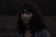 一位黑发蓬乱的年轻女性，站在自然环境中，背景模糊，目光闪烁地看着镜头