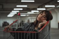 Dreamy female with black hair and head tilted to side with hand near face sitting in metal cart while looking at camera on blurred underground parking lot