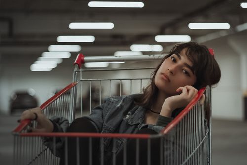 Free Dreamy female with black hair and head tilted to side with hand near face sitting in metal cart while looking at camera on blurred underground parking lot Stock Photo