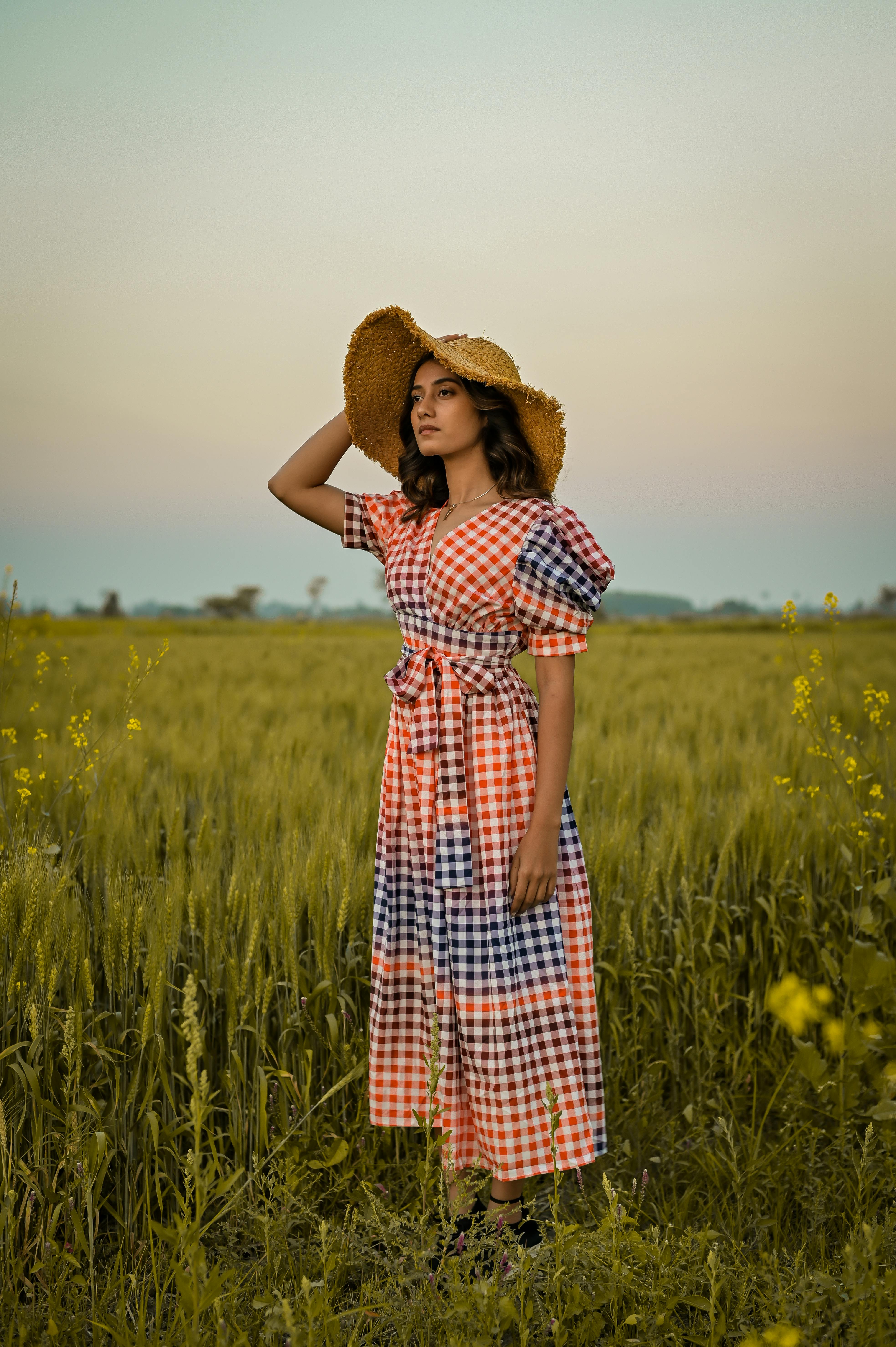 Young woman in a checkered dress and straw hat standing in a serene field during sunset.