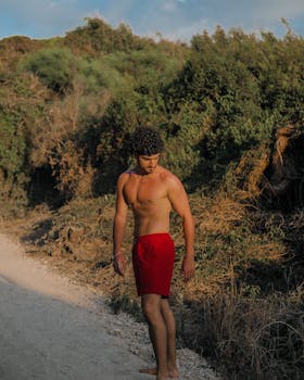 A shirtless man with curly hair walks on a sunny path in Paestum, Italy, showcasing natural beauty.