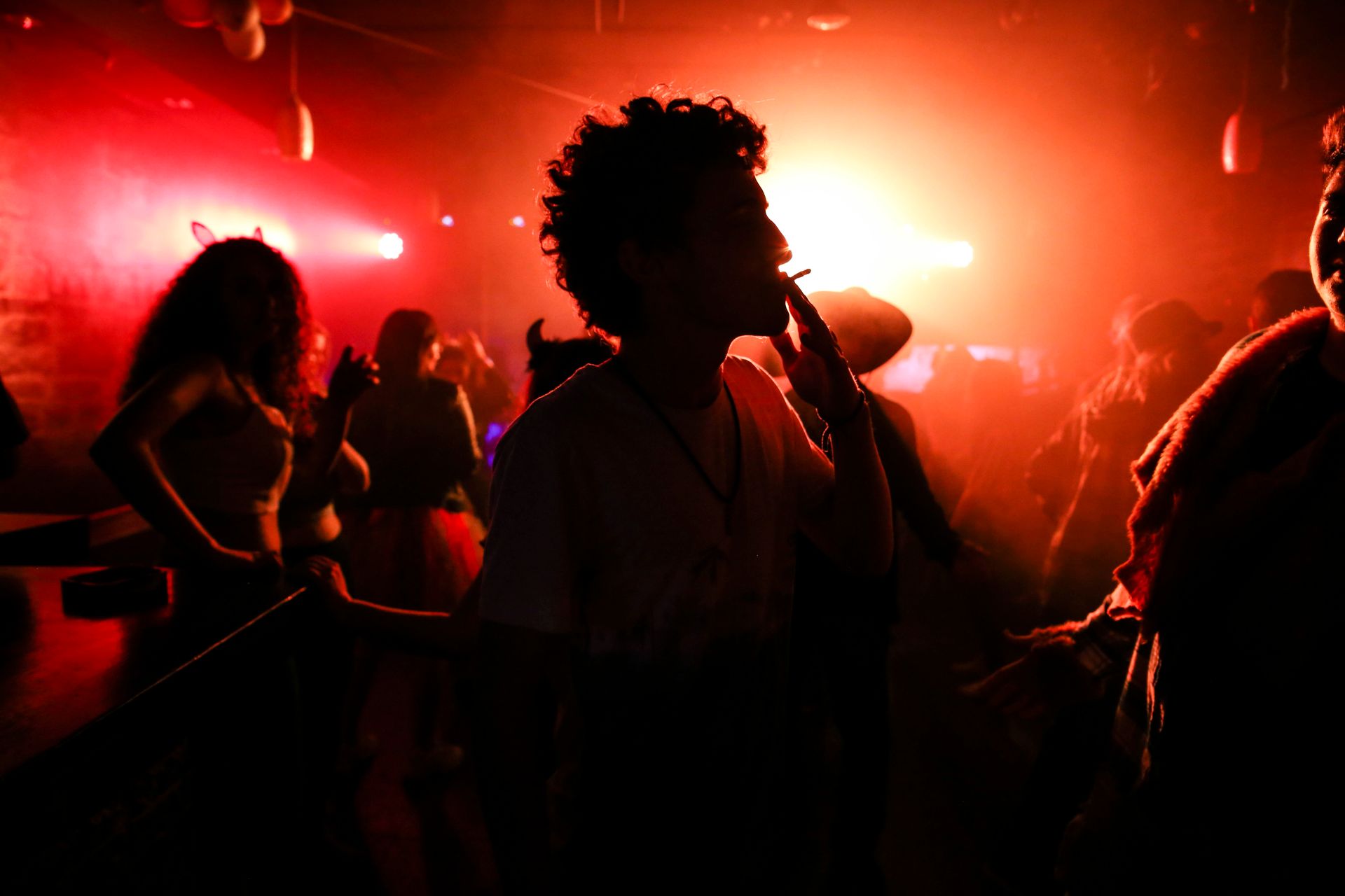 Silhouetted nightlife scene with people dancing and smoking in red lighting.