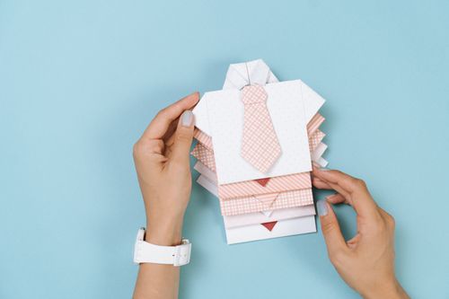 Free A stylish origami shirt with tie held by hands, displayed on a blue surface. Perfect for design inspiration. Stock Photo