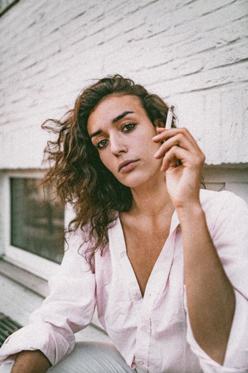 Free A contemplative woman with wavy hair smokes while posing in a pink shirt outdoors. Stock Photo