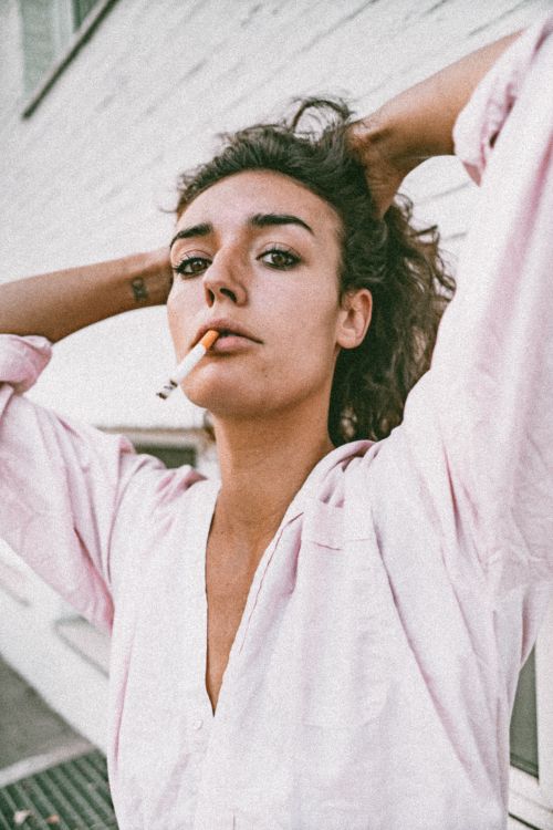Free Woman with cigarette outside, wearing pink sleeves. Urban vibe, relaxed elegance in Brussels. Stock Photo