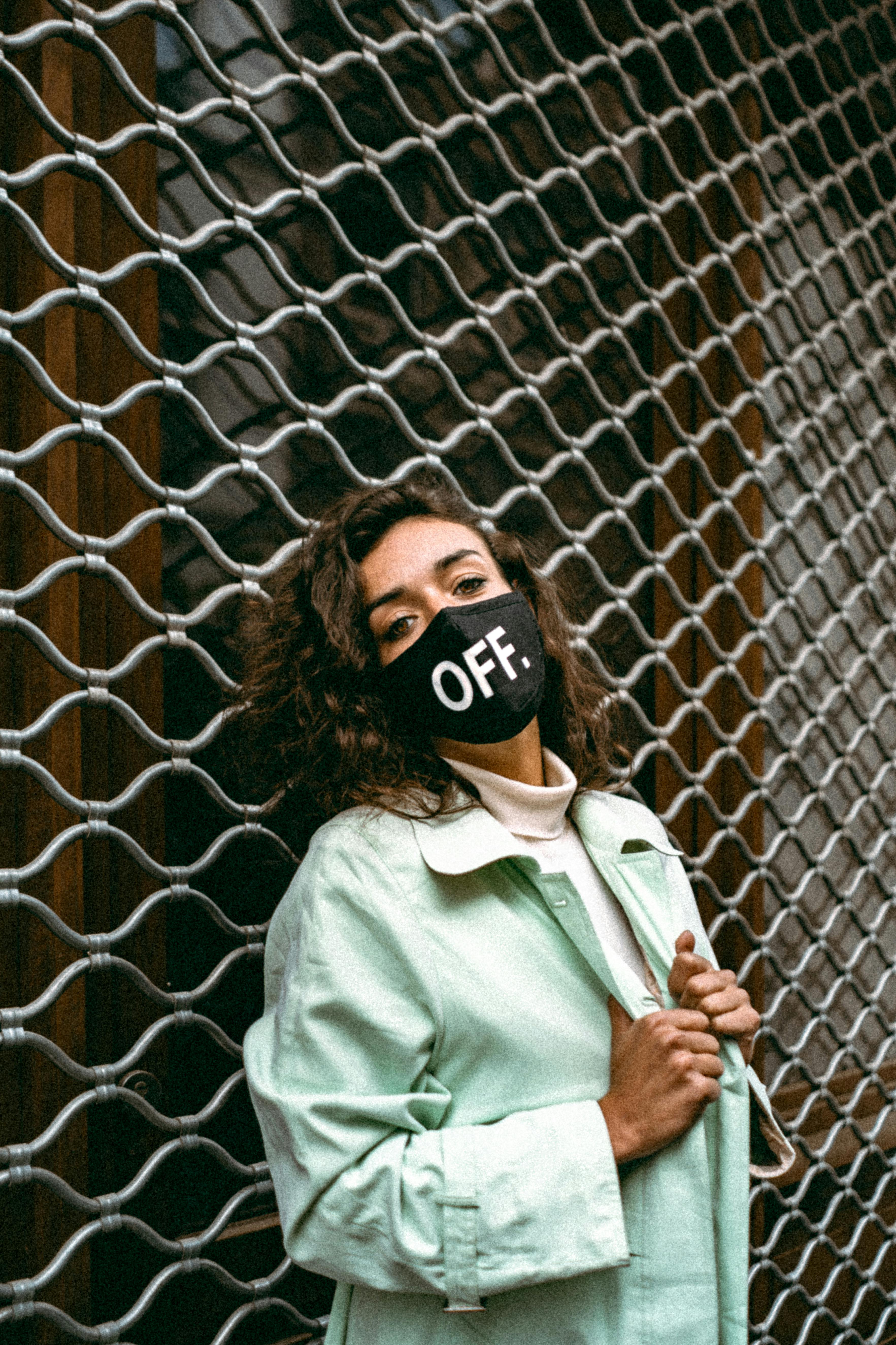 Stylish woman in green coat and face mask posing outdoors by a metal fence.