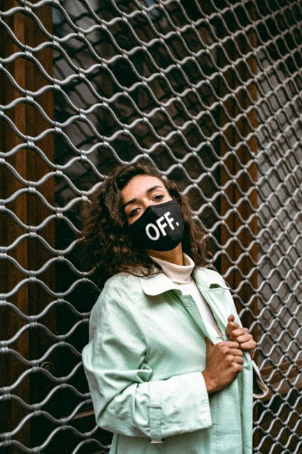 Stylish woman in green coat and face mask posing outdoors by a metal fence.