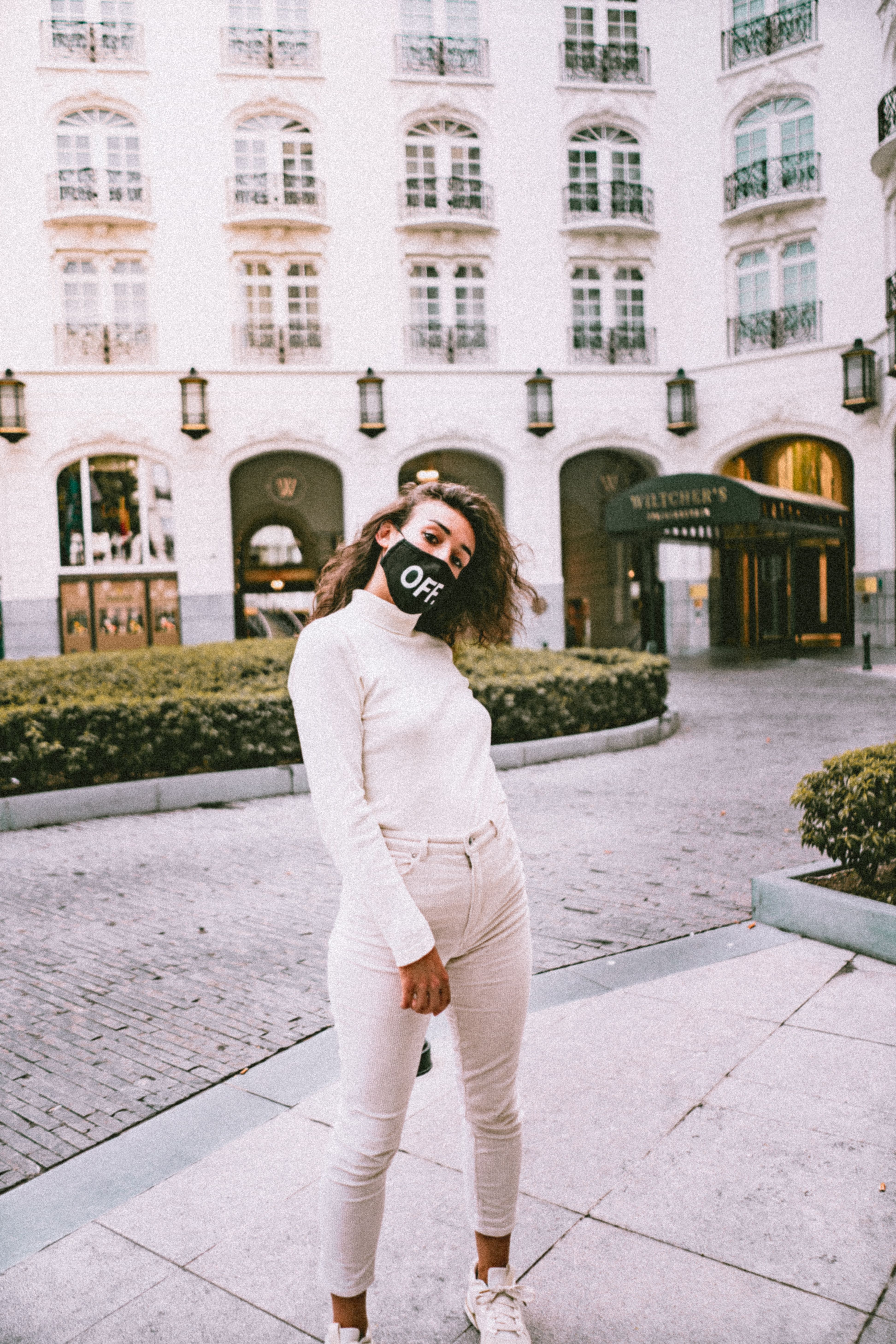 A woman in a turtleneck and mask stands outside a classic European hotel in Brussels.