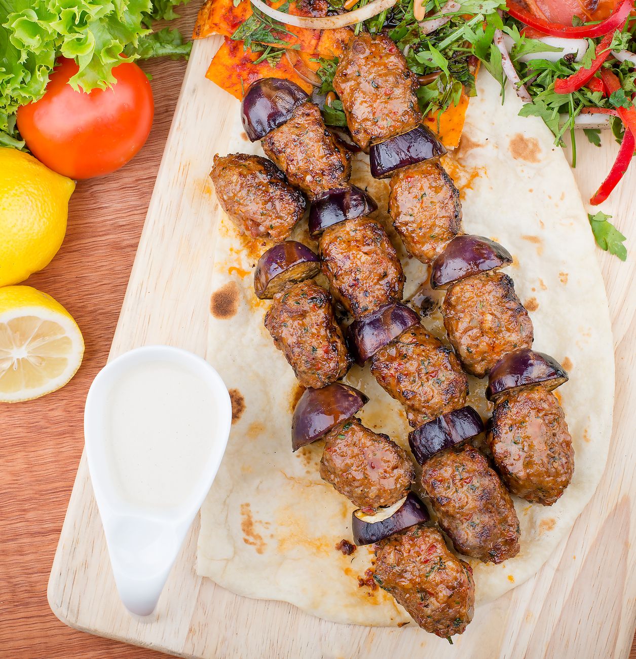 Four grilled adana kebabs sliding off metal skewers onto warm flatbread, topped with fresh parsley, sliced red onion, and lemon wedges, showing dark char on the exterior.