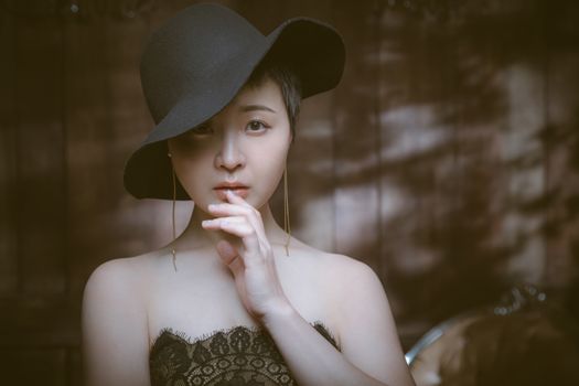 Young dreamy Asian woman in stylish clothes touching lips while looking at camera in house