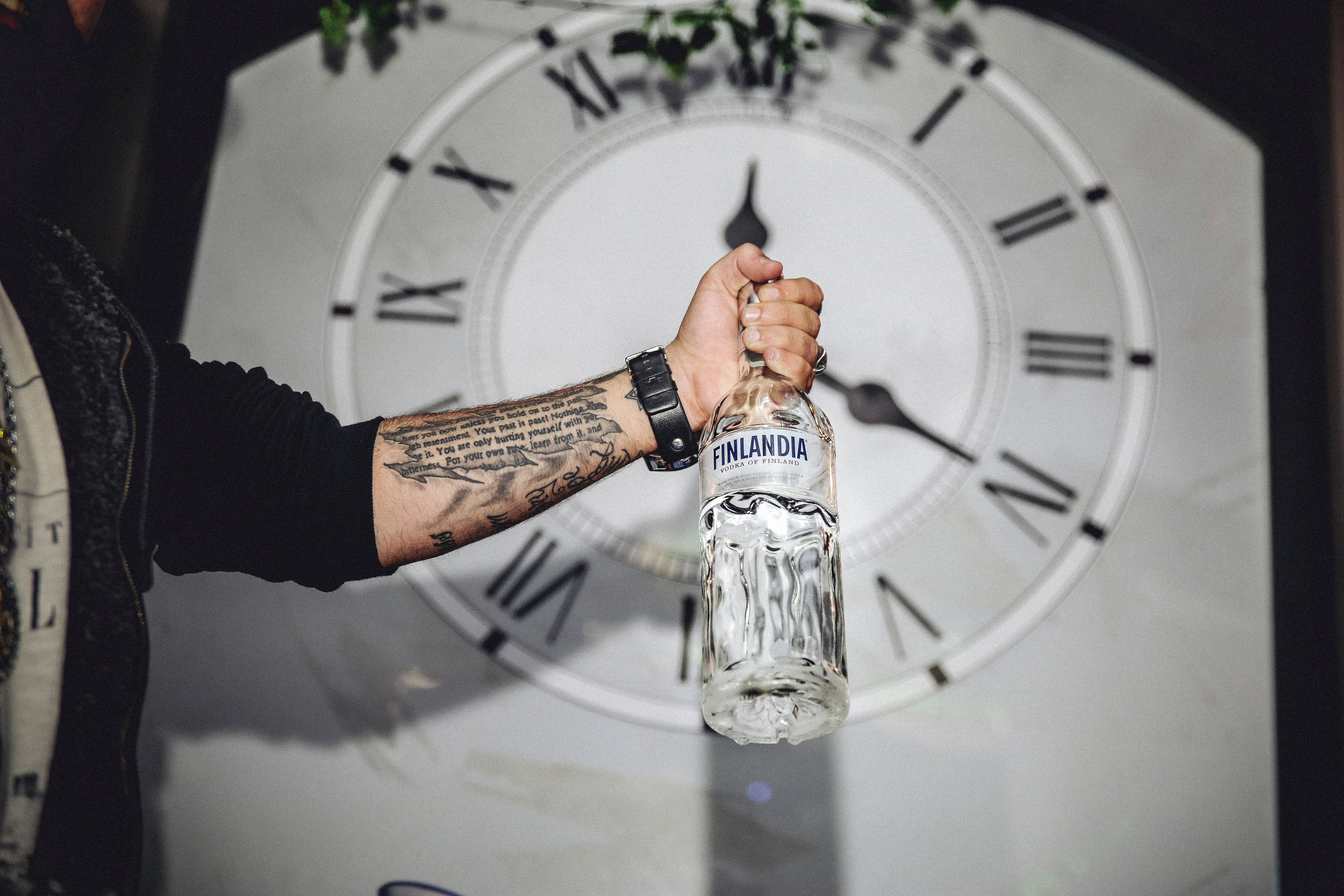 A tattooed arm holding a Finlandia vodka bottle in front of a large clock face.