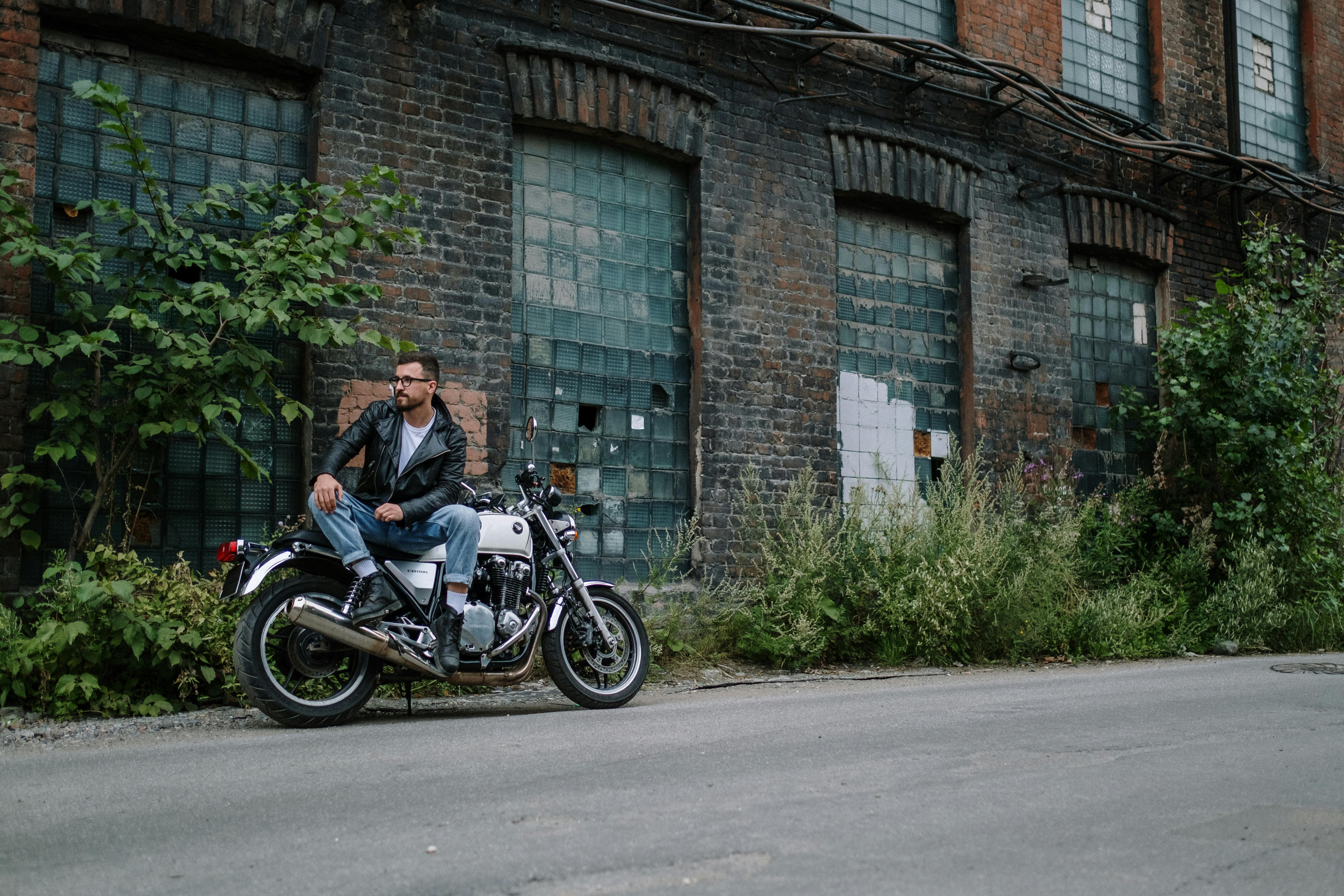 A man in a leather jacket sits on his motorcycle beside an abandoned building, exuding a cool and adventurous vibe.