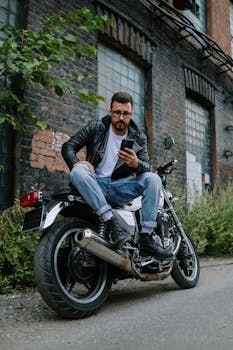 A man in a leather jacket sits on a motorcycle using a smartphone in an industrial setting.