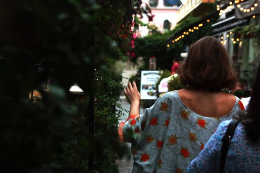 Back view of a woman with short hair walking through a lively, plant-filled alleyway adorned with lights.