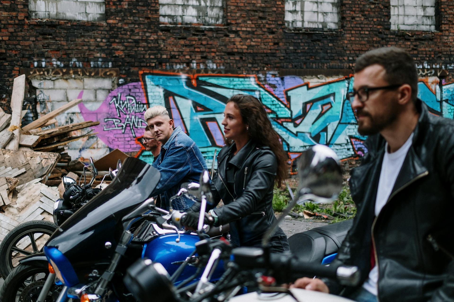 Three bikers with motorcycles against a backdrop of urban graffiti and a gritty brick wall.