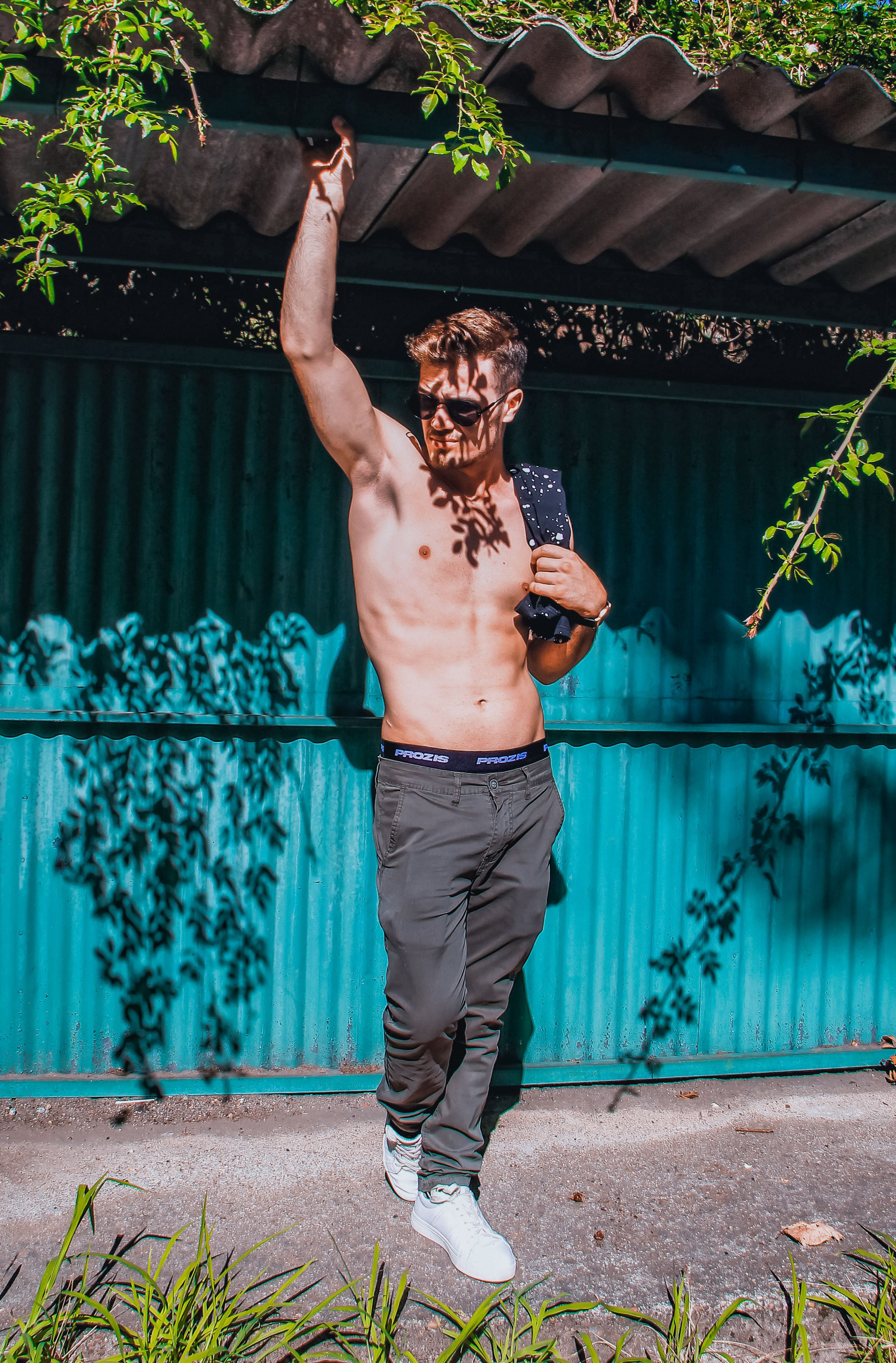 Muscular shirtless man with sunglasses posing outdoors under dappled sunlight.