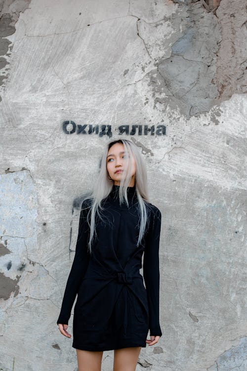 Beautiful young Asian slender female in black dress with long blond straight hair thoughtfully looking away while standing by old dirty shabby wall in daylight