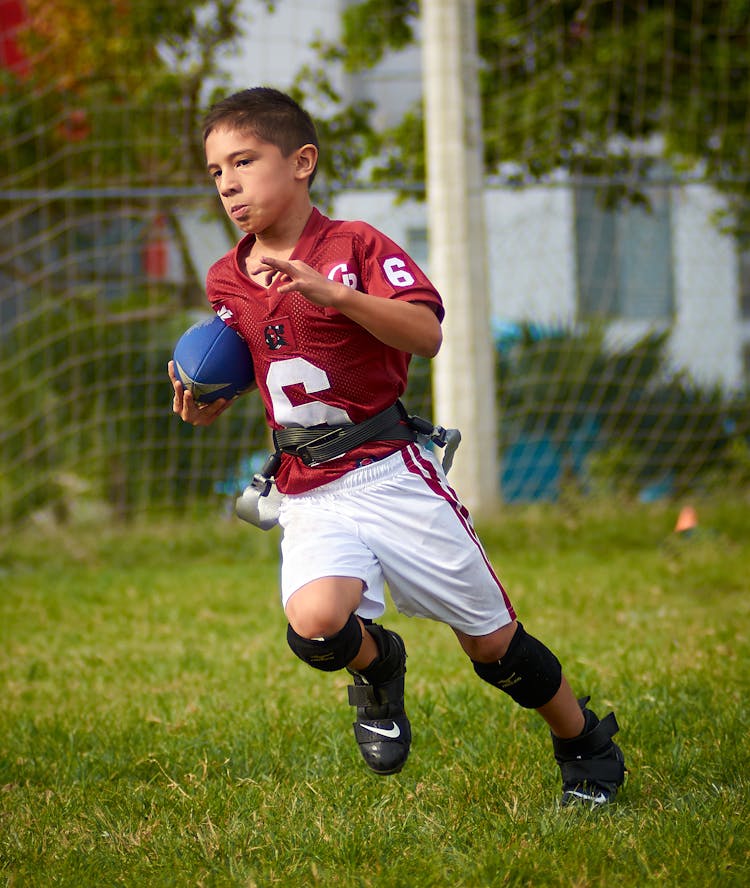 Young Boy In Red And White Sports Uniform Running With The Ball Playing American Football