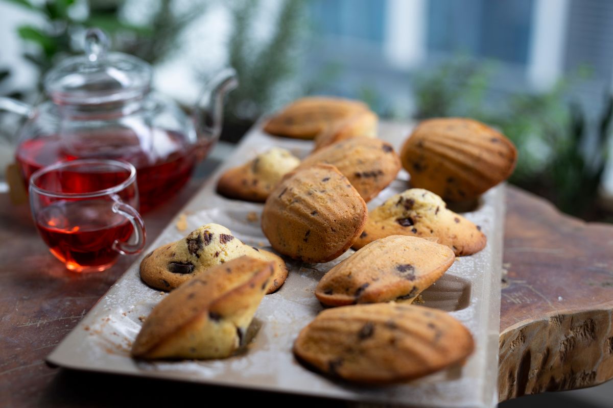 Madeleines with chocolate chips in a baking tray beside a glass teapot of red tea