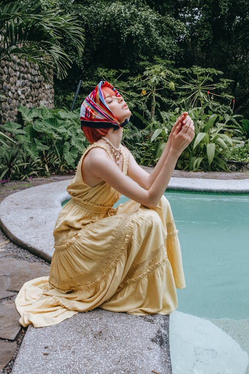 Side view full length charming young female in yellow maxi dress and silk headscarf sitting near swimming pool in tropical resort