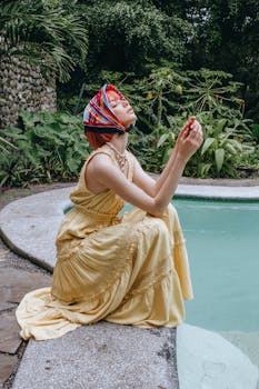 Side view full length charming young female in yellow maxi dress and silk headscarf sitting near swimming pool in tropical resort
