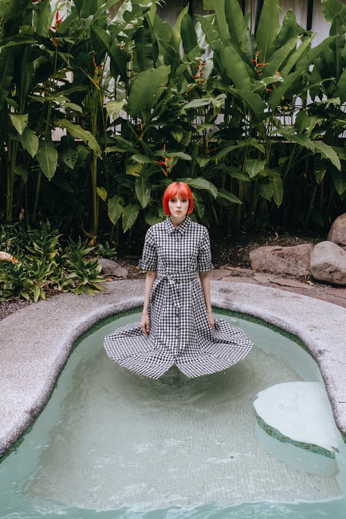 Phlegmatic young female in gray dress standing calmly in swimming pool with skirt floating on water surface in exotic resort