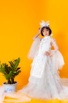 一位身着优雅白色连衣裙、头戴帽子的年轻女孩，在充满活力的橙色背景前摆出姿势，身旁有一棵盆栽，整体散发着时尚与风格