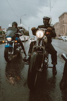 Motorcyclists riding through a city street on a rainy day with slick roads and traffic.