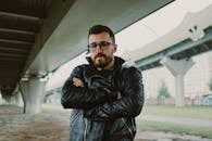 一名戴眼镜、留着胡子的男子在城市桥下穿着皮夹克摆姿势