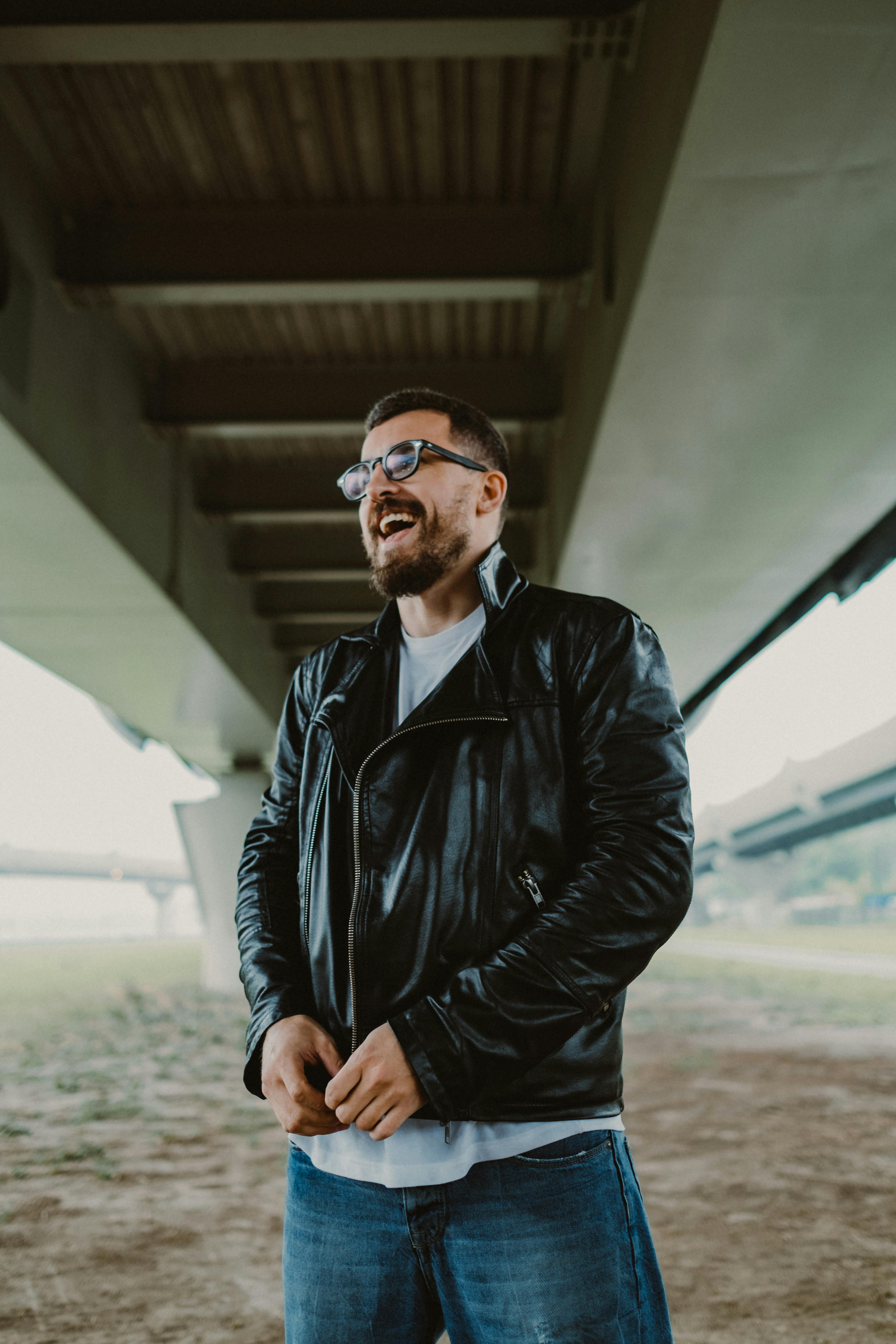 桥下站着一名身穿黑色皮夹克、戴着眼镜的开朗男子，他笑容满面