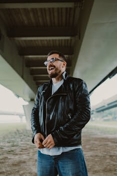 桥下站着一名身穿黑色皮夹克、戴着眼镜的开朗男子，他笑容满面