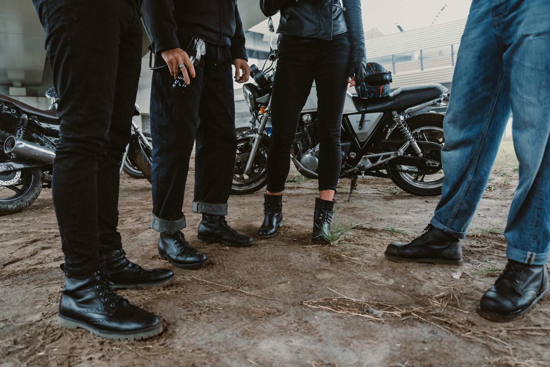 A group of bikers in jeans and leather standing together with motorcycles outdoors.