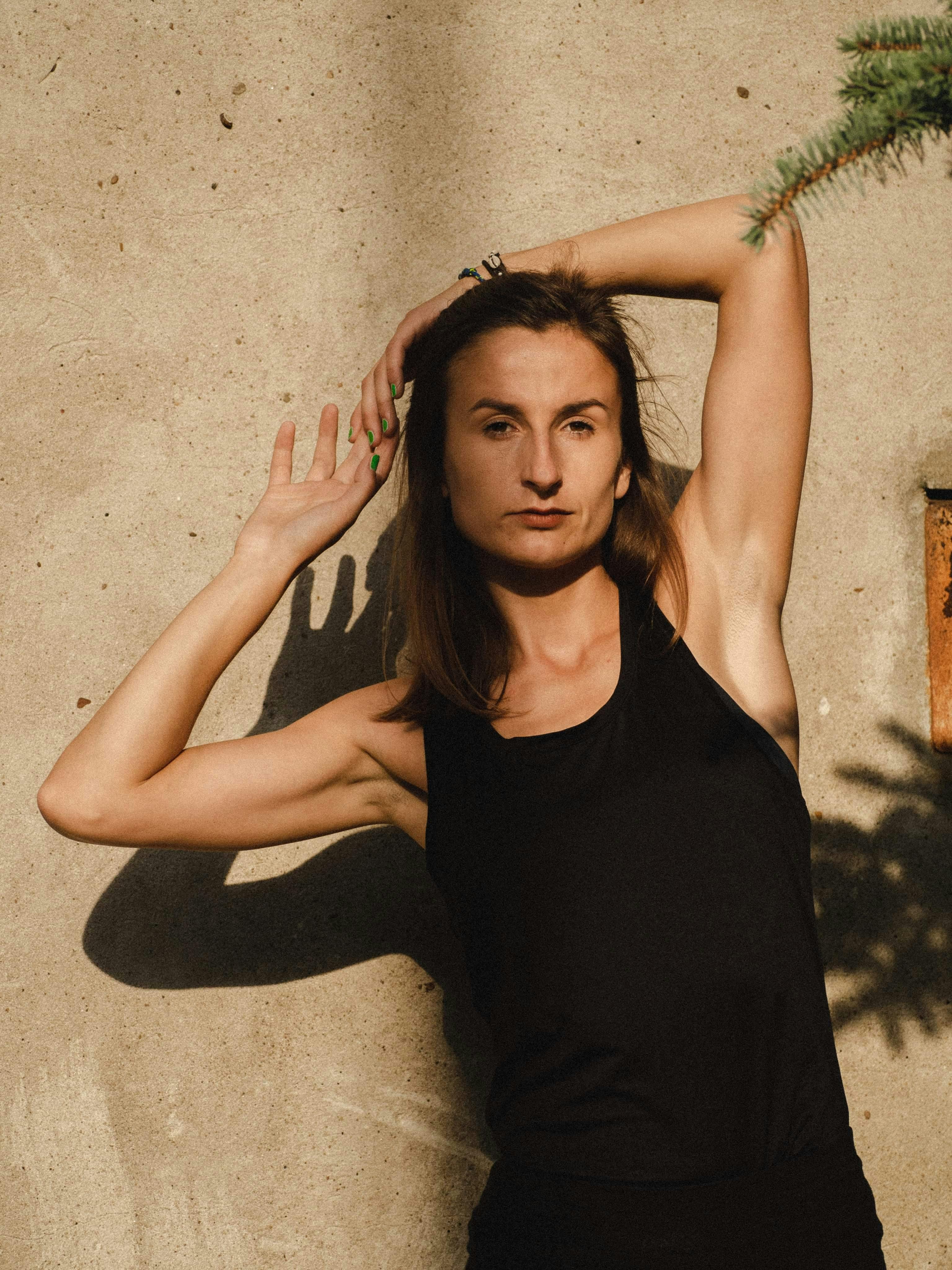 A woman in a black tank top poses gracefully against a sunlit wall, exuding beauty and confidence.