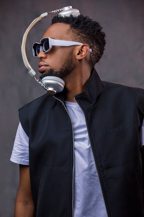 Young African American man with curly hair and beard in trendy clothes and sunglasses standing with headphones in studio